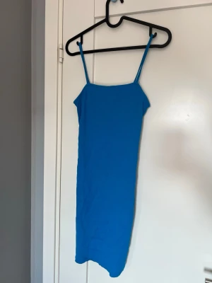 Blå tight klänning med smala axelband - En enkel och stilren blå klänning med smala axelband och rak passform. Klänningen är kort  och sydd i ett stretchigt tyg som sitter tajt mot kroppen. Perfekt för dig som gillar minimalistisk stil och vill ha något bekvämt men snyggt.