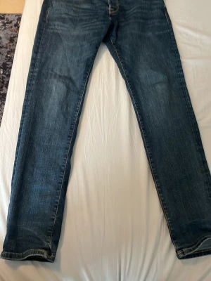 Blå jeans från Jack & Jones - Snygga blå jeans från Jack & Jones med klassisk femficksmodell och raka ben. Jeansen har slitningar och kontrastsömmar som ger en cool look. Tillverkade i denim med bälteshällor och knappgylf. Perfekta för en avslappnad stil.