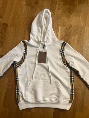 Vit Burberry hoodie med rutiga detaljer - Säljer en vit hoodie från Burberry med klassiska beige, svarta och röda rutiga detaljer längs sidorna och axlarna. Hoodien har stor huva, dragsko och en stor magficka. Tillverkad i mjukt bomullsmaterial för en skön och avslappnad look.