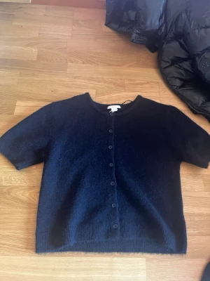 Mörkblå kortärmad kofta - Snygg mörkblå kofta med korta ärmar och rund halsringning. Koftan har knäppning framtill med små knappar och är gjord i ett mjukt, fluffigt material som ger en mysig känsla. Perfekt att slänga över en topp för en chill look.