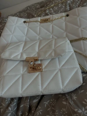 Vit quiltad handväska med guldkedja - 2at Snygga vita handväskor i quiltat mönster med guldfärgad kedja och låsdetalj framtill. Väskan har en klassisk rektangulär form och lockstängning. Perfekt för dig som gillar stilrena accessoarer med en touch av lyx.❤️