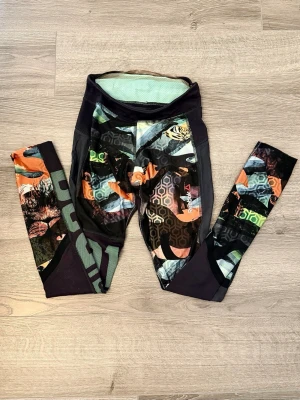 Färgglada tights från Reebok - Unikt mönster med geometriska former (hexagoner), kamouflage och färgstarka detaljer. Tightsen har mesh på sidorna med god stretch.   Storlek är jag osäker på, men den skulle troligtvis passa XXS eller för barn 10-12yrs. Därför är priset satt utefter detta. Vill bara bli av med dessa!