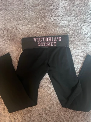 Svarta leggings Victoria's Secret - Säljer ett par svarta leggings från Victoria's Secret med bred grå midjeresår. På baksidan står det 'VICTORIA'S SECRET' i vitt och rosa och på baksidan finns ett litet tryck med änglavingar. Supersköna och perfekta till alla outfits. Säljer då de tyvärr är lite för korta🤍Superrrr sköna