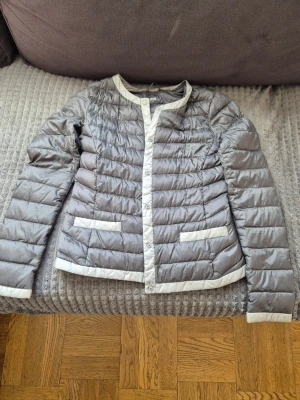 Grå pufferjacka med vita detaljer - Snygg grå pufferjacka med vita kontrastdetaljer längs kanter, muddar och fickor. Jackan har rund halsringning, tryckknappar framtill och två fickor. Quiltad design och lätt vadderad för extra värme. Perfekt för kalla dagar.