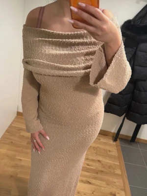 Beige stickad offshoulder långklänning - Elegant beige långklänning med offshoulder-design och vida ärmar. Klänningen har en glittrig textur och smickrande passform som framhäver figuren. Perfekt för speciella tillfällen när du vill sticka ut.