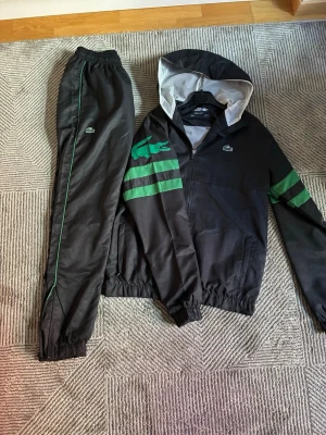 Svart träningsset från Lacoste - Snyggt svart träningsset från Lacoste med grön logga och gröna ränder på jackans ärmar. Setet består av en zip-hoodie med huva och matchande byxor med resår i midja och benslut. Materialet är lätt och sportigt, perfekt för träning eller chill. Klassisk Lacoste-stil med krokodillogga. 
