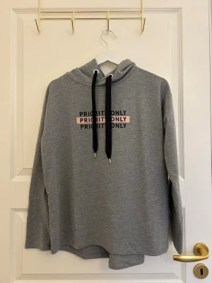 Grå hoodie med tryck och huva - Snygg grå hoodie med svart text 'PRIORITY ONLY' tryckt tre gånger på bröstet och en ljusrosa bakgrundsremsa. Hoodien har svarta dragsnören i huvan och är tillverkad i mjukt sweatshirtmaterial. Perfekt för en chill och avslappnad stil.