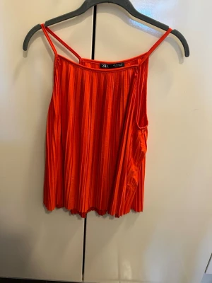 Plisserad orange/röd blus från Zara - Trendig orange plisserad blus från Zara med tunna axelband och lös passform. Blusen har ett snyggt fall och är tillverkad i ett lätt, glansigt tyg som ger en cool look. Perfekt för dig som vill sticka ut med färg och stil.
