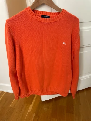 Vintage stickad tröja från Burberry - Säljer en vintage orange stickad tröja från Burberry London. Tröjan har rund halsringning, ribbade muddar och en liten vit Burberry-logga broderad på bröstet. Tillverkad i Italien och perfekt för dig som gillar klassisk men färgstark stil. Strl. L men sitter mer som M! 