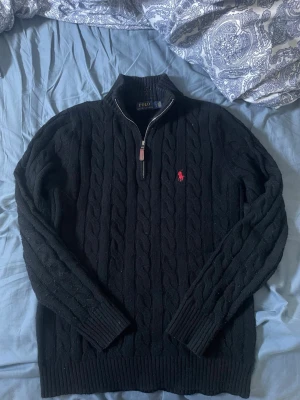 Svart kabelstickad halfzip Polo Ralph Lauren - Kabelstickad svart tröja från Polo Ralph Lauren med halv dragkedja och röd broderad logga på bröstet. Tröjan har ribbade muddar och hög krage, perfekt för lager på lager. Klassisk och stilren design i mjukt material.