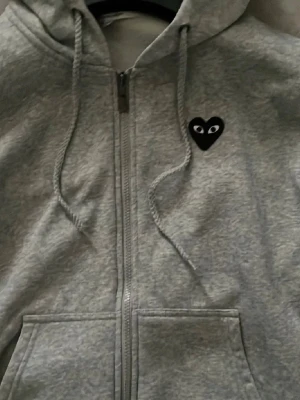 CDG Play Zip Hoodie Comme Des Garcons - Säljer en grå zip-hoodie från Comme des Garçons Play med det ikoniska svarta hjärtat med ögon på bröstet och en stor version på ryggen. Hoodien har dragkedja, huva med snören och två fickor framtill. Materialet är mjukt och bekvämt