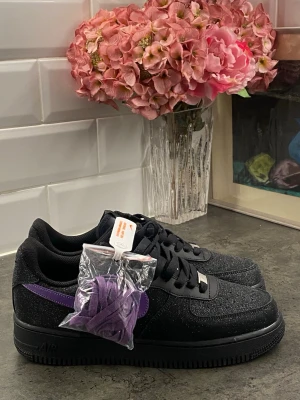 Nike Air Force 1 svart/lila sneakers - Snygga Nike Air Force 1 sneakers i svart med lila swoosh och glitterdetaljer. Skorna har en klassisk låg siluett, svart yttersula och kommer med extra lila skosnören. Perfekta för dig som vill sticka ut med stilrena men unika sneakers.