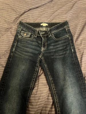 Mörkblå bootcut jeans  - Snygga mörkblå bootcut jeans med slitningar och tydliga kontrastsömmar. Klassisk femficksmodell med dekorativa bakfickor och knappdetaljer. Jeansen har låg midja och är tillverkade i denim med lätt stretch för skön passform.