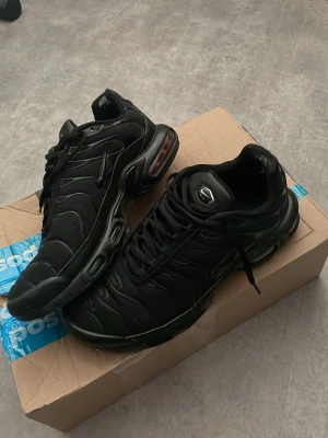 Nike Air Max Plus helsvarta sneakers - Säljer ett par helsvarta Nike Air Max Plus sneakers med klassisk TN-logga på plösen. Skorna har mesh- och syntetöverdel, synlig Air Max-dämpning i sulan och svarta snören. Snygg streetstil och ikonisk design med vågiga linjer på sidan.              Använt flera gånger, syns skicken på skorna i bilderna!!!!!