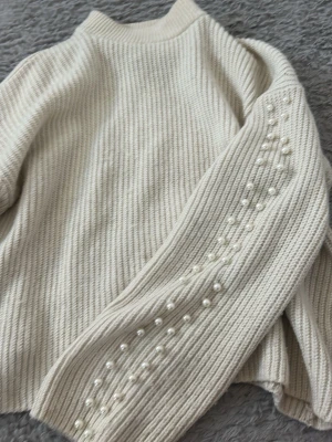 Beige stickad tröja med pärlor - Snygg beige stickad tröja med ribbad struktur och pärldetaljer längs ärmarna. Tröjan har hög hals och långa ärmar, perfekt för dig som gillar en trendig och elegant look. Materialet känns mjukt och skönt mot huden.