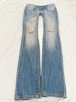 Ljusblå bootcut jeans med slitningar - Säljer ett par ljusblå bootcut jeans med slitna detaljer på låren och knäna. Jeansen har klassisk femficksmodell, låg midja och snyggt tvättad denim. Perfekta för en avslappnad och trendig look.