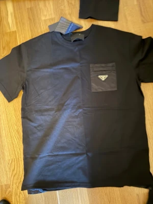 Svart t-shirt från Prada med ficka - Snygg svart t-shirt från Prada med rund halsringning och en bröstficka med ikonisk Prada-logga i metall. Tillverkad i mjukt bomullstyg och har en clean, minimalistisk look som passar perfekt till jeans eller shorts.