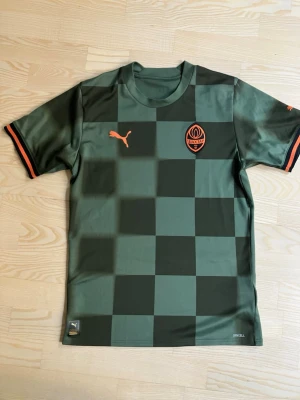 Grön Shakhtar Donetsk matchtröja Puma - Snygg grön matchtröja från Puma med Shakhtar Donetsk-logga på bröstet. Tröjan har rutigt mönster i olika gröna nyanser och orange detaljer på ärmar och loggor. Tillverkad i ett lätt och funktionellt material som passar perfekt till träning eller match.