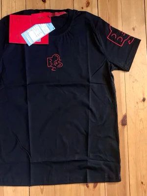 Svart t-shirt från Hugo Boss - Svart t-shirt från Hugo Boss med röd broderad logga på bröstet och ärmen. Klassisk rund halsringning och korta ärmar. Tillverkad i mjuk bomull som känns skön mot huden. Perfekt för en stilren och modern look.