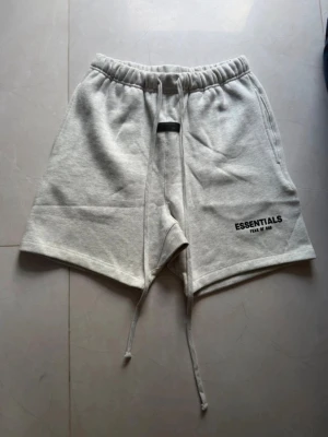 Essentials Fear of God shorts  - Grå sweatshorts från Essentials Fear of God med elastisk midja och långa snören framtill. Klassisk logga tryckt på vänster ben. Mjuk bomullskänsla och avslappnad passform, perfekta för chill dagar.