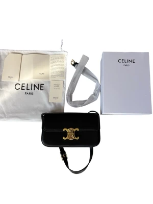 Svart Triomphe handväska från Celine - Snygg svart handväska från Celine med ikoniskt Triomphe-spänne i guld framtill. Väskan är tillverkad i slätt läder och har en justerbar axelrem. Kommer med original dustbag, box och certifikat. Perfekt för dig som gillar lyxiga accessoarer med stilren design.