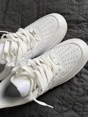 Nike Air Force 1 vita sneakers - Klassiska Nike Air Force 1 sneakers i helvitt läder med perforerad tåbox, tjock vit sula och vita skosnören. Ikonisk design med Nike-logga på sidan och metallbricka på snörningen. Perfekta för en clean och stilren look.