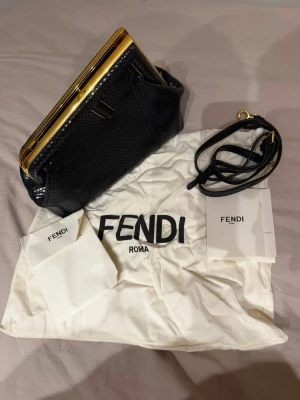 Svart kuvertväska i ormskinn från Fendi - Snygg svart kuvertväska från Fendi i ormskinnsmönstrat läder med guldfärgade metalldetaljer. Väskan har avtagbar axelrem och ikoniskt Fendi-mönstrat tygfoder. Kommer med dustbag och äkthetskort. Perfekt för dig som vill ha något lyxigt och trendigt.