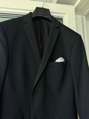 Svart kavaj  - Stilren svart kavaj med klassisk passform och spetsiga slag. Kavajen har en vit näsduk i bröstfickan och två fickor framtill. Perfekt för dig som vill ha en clean och tidlös look.