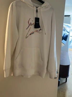 Vit Saint Laurent hoodie med tryck - Säljer en vit hoodie från Saint Laurent med färgglatt logotyptryck på bröstet. Hoodien har huva med dragsko och en stor magficka framtill. Perfekt för dig som gillar stilrena och exklusiva plagg med en twist. Litet märke på den just nu men går bort i tvätten 