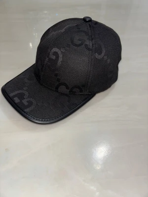 Svart keps från Gucci med GG-mönster - Snygg svart keps från Gucci med klassiskt GG-monogram i ton-i-ton. Kepsen har böjd skärm med läderdetalj och justerbar rem baktill. Tillverkad i textilmaterial som ger en exklusiv känsla och passar perfekt till streetwear eller en trendig look.