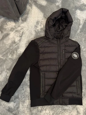 Svart pufferjacka Storlek M - Säljer en svart pufferjacka från Suply and demand med quiltad front och huva. Jackan har mjuka ärmar och rygg i stretchigt material, dragkedja framtill och en Superdry-logga på vänster ärm. Perfekt för kalla vinterdagar. Storlek M