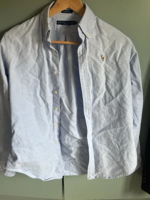 Ljusblå skjorta från Ralph Lauren - Klassisk ljusblå skjorta från Ralph Lauren med button-down krage och broderad logga på bröstet. Skjortan har långa ärmar, vita knappar och är tillverkad i mjuk bomull. Perfekt för en clean och stilren look. Notera att den är i junior-storlek