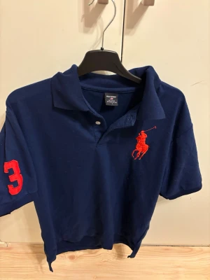 Marinblå pikétröja från Ralph Lauren - Marinblå pikétröja från Ralph Lauren med röd broderad logga på bröstet och en stor röd siffra 3 på ärmen. Klassisk krage och korta ärmar. Perfekt för en sportig och stilren look.