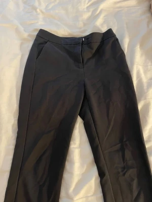 Svarta kostymbyxor med fickor - Snygga svarta kostymbyxor med raka ben och diskreta sidofickor. Byxorna har en klassisk midja och dragkedja framtill. Perfekta för en clean och stilren look. Materialet ser ut att vara en mjuk och lite stretchig bomullsblandning.