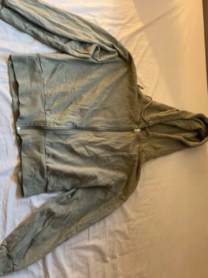 Grön hoodie från 157 - Säljer en grön hoodie från 157 med dragkedja framtill och justerbar huva. Hoodien har långa ärmar och breda muddar vid ärmslut och nederkant. Tillverkad i mjukt bomullsmaterial som är skönt att ha på sig. Perfekt för en avslappnad stil.