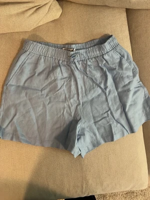 Ljusblå linneshorts från NA-KD - Säljer ett par ljusblå shorts från NA-KD i linne med avslappnad passform. Shortsen har elastisk midja med snörning och sidofickor. Perfekta för varma dagar och enkel att matcha med olika toppar.