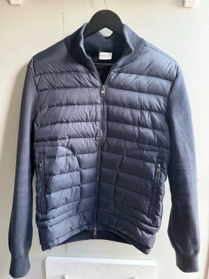 Mörkblå cardiganjacka från Moncler - Snygg mörkblå cardiganjacka från Moncler med quiltad front och stickade ärmar. Jackan har dragkedja framtill, två sidofickor med dragkedja och en liten Moncler-logga på ärmen. Perfekt för lager-på-lager och stilren look.