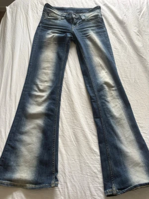Blå bootcut jeans med slitningar - Säljer ett par blå bootcut jeans med tydliga slitningar och ljusa partier längs benen. Jeansen har klassisk femficksmodell och låg midja. Materialet är denim i bomull med lite stretch för extra komfort. Perfekta för en avslappnad och trendig look.