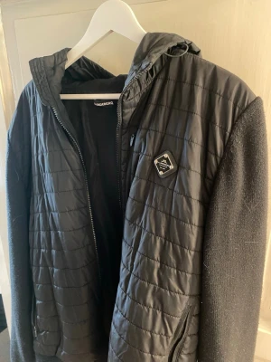 Svart cardigan från J.Lindeberg - Snygg svart vindjacka från J.Lindeberg med quiltad front, huva och ribbade muddar. Jackan har dragkedja framtill och en diskret logotyp på bröstet. Passar M och L. 