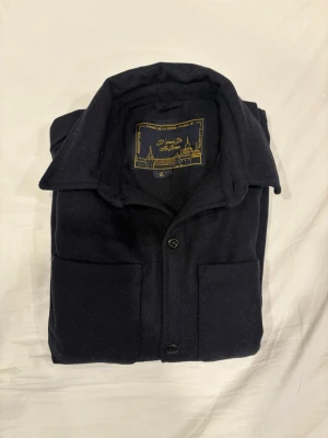 D’once de la sense overshirt mörkblå - Stilren mörkblå overshirt från D’once de la sense med klassisk krage och två stora bröstfickor. Skjortjackan har knäppning framtill och är tillverkad i ett mjukt, tjockt tyg i 100% ull som ger en clean och modern look. Perfekt att bära lager på lager under kyligare dagar. 