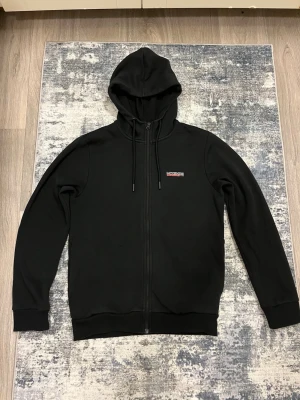 MCKENZIE - Svart hoodie med dragkedja från Napapijri. Klassisk modell med huva och justerbara snören, samt liten logotyp på bröstet. Perfekt för en avslappnad och stilren look.