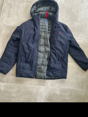 Mörkblå pufferjacka från Polo Ralph Lauren - Snygg mörkblå pufferjacka från Polo Ralph Lauren med röd logga på bröstet. Jackan har huva, dragkedja och två fickor med dragkedja. Insidan är quiltad i grått material för extra värme. Perfekt för kalla vinterdagar. Nypris 4800 säljer nu för 1800