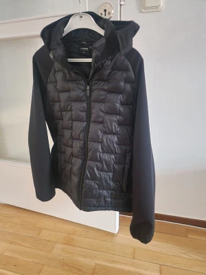 Svart pufferjacka från Everest - Snygg svart pufferjacka från Everest med huva och dragkedja framtill. Jackan har quiltad front och rygg i glansigt material samt släta ärmar. Perfekt för kalla vinterdagar och har en sportig look.
