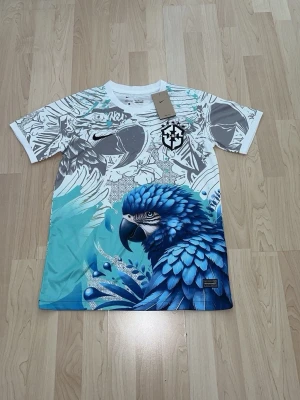 Nike t-shirt med blå papegojtryck - Unik t-shirt från Nike med stort blått papegojmotiv framtill och detaljer i vitt, grått och turkost. Kortärmad modell med rund hals och tryckt Nike-logga på bröstet. Perfekt för dig som vill sticka ut med en färgstark och kreativ stil.