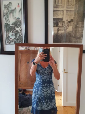 Blå blommig sommarklänning - Söt blå klänning med vitt blommönster och v-ringning. Klänningen har korta ärmar med spetsdetaljer och smock i ryggen för skön passform. Perfekt för varma dagar och har en feminin, figurnära siluett.