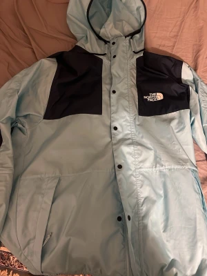 The North Face Jacka - Snygg ljusblå vindjacka från The North Face med svarta paneler på axlar och bröst. Köpt från JD-Sports för 1250kr Jackan har huva, dragkedja och tryckknappar framtill samt två stora fickor. Perfekt för blåsiga dagar och streetwear-stil.