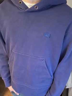 Blå hoodie från Acne Studios - Säljer en blå hoodie från Acne Studios med klassisk känguruficka och den ikoniska lilla ansiktsloggan på bröstet. Hoodien har huva med dragsko och ribbade muddar vid ärmar och nederkant. Perfekt för en chill och stilren look.