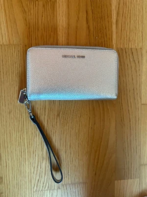 Silvrig plånbok från Michael Kors - Snygg silvrig plånbok från Michael Kors med avtagbar handledsrem. Plånboken har en stilren, rektangulär form och är tillverkad i ett glansigt, strukturerat material. Perfekt för att hålla koll på kort och cash med en touch av lyx.