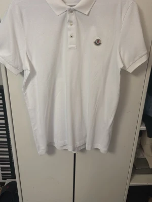 Vit Moncler pikétröja med logga - Klassisk vit pikétröja från Moncler med broderad logga på bröstet. Tröjan har krage, knäppning med tre knappar och korta ärmar. Perfekt för en clean och stilren look. Loggan är tyvärr utstrechad lite pga min katt som klösde på den men jag tror det går att fixa med hjälp av ett stryk gärn, därav priset. Men det är inget som märks tydligt.