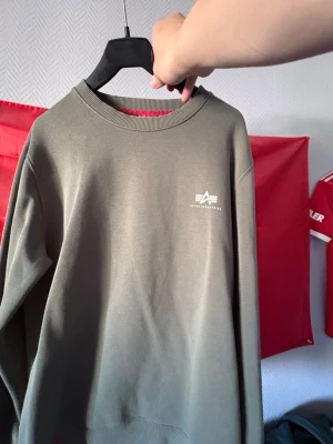 Olivgrön sweatshirt från Alpha Industries - Snygg olivgrön sweatshirt från Alpha Industries med rund halsringning och diskret logga tryckt i vitt på bröstet. Tröjan har ribbade muddar vid ärmslut och nederkant. Perfekt för en avslappnad och stilren look.
